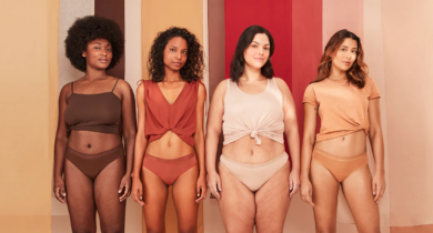 Quatro pessoas posam lado a lado diante de um fundo em tons terrosos e rosados. Elas vestem roupas íntimas e tops em cores neutras e quentes, com diferentes estilos de corpo e tons de pele, representando diversidade e autoestima. Todas olham para a câmera com expressões serenas e confiantes.
