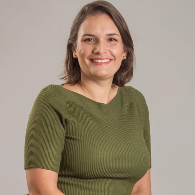 Foto mostra mulher branca de cabelos curtos e castanhos, com camisa de manga verde, em background branco. Ela sorri para a foto.